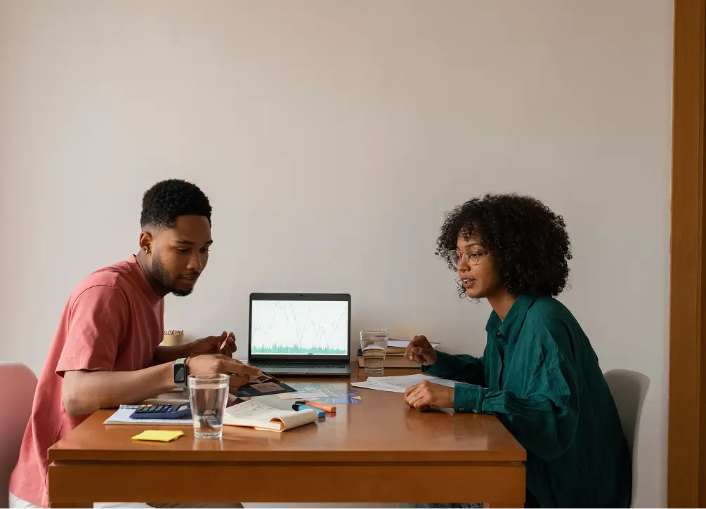 two people sitting at a table with a laptop and papers
