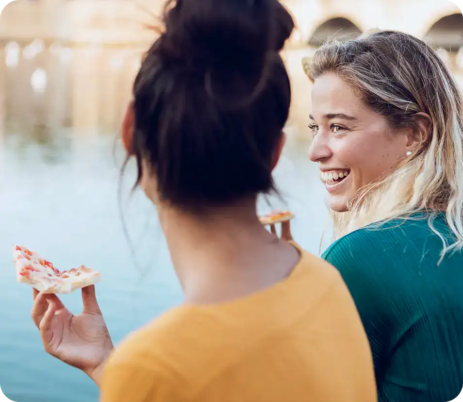 there are two women that are eating pizza together by the water